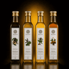 Four glass bottles of Kaizer cold-pressed oils arranged in a row on a reflective dark surface against a black background with a warm golden glow behind them. The bottles contain olive oil in varying shades from dark green to amber-brown, each with a white label showing an illustration of its respective variety: avocado, green olives, garlic, and peanuts.